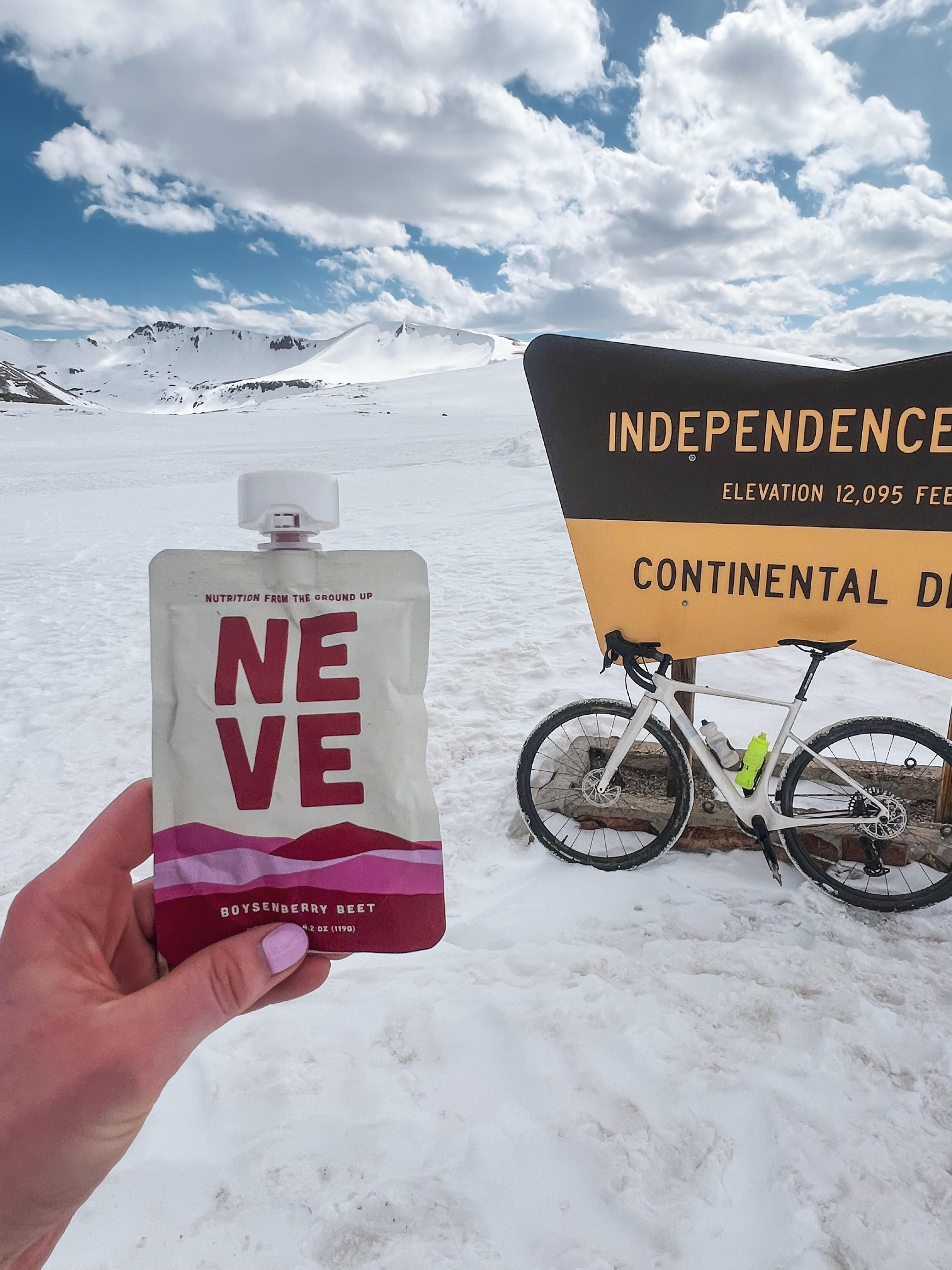 Hand holding a Neve Boysenberry Beet smoothie pouch with a snowy mountain landscape and bike in the background on Independence Pass in Colorado.