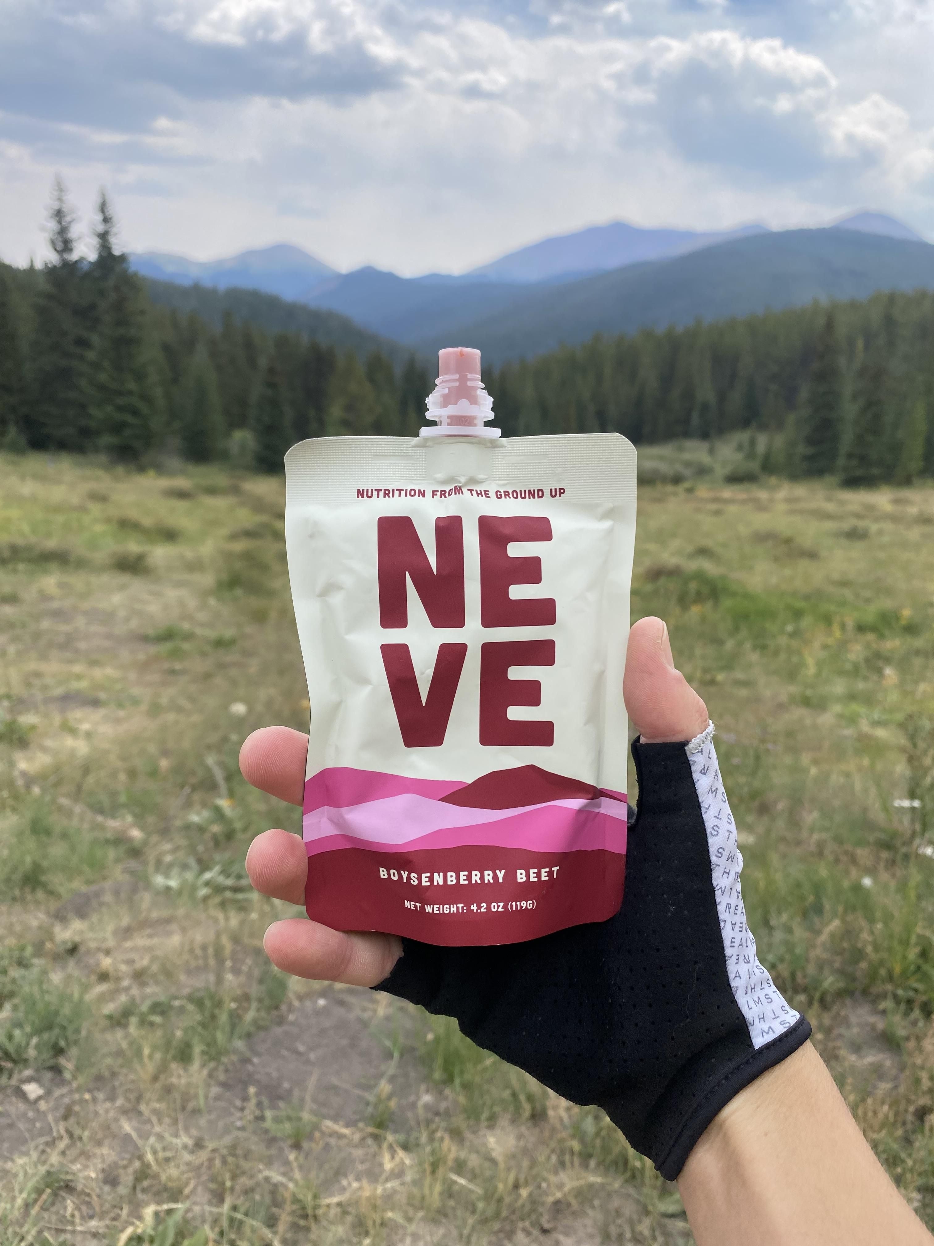 Hand wearing a cycling glove holding a Neve Boysenberry Beet smoothie pouch, with a mountain meadow and pine forest in the background.