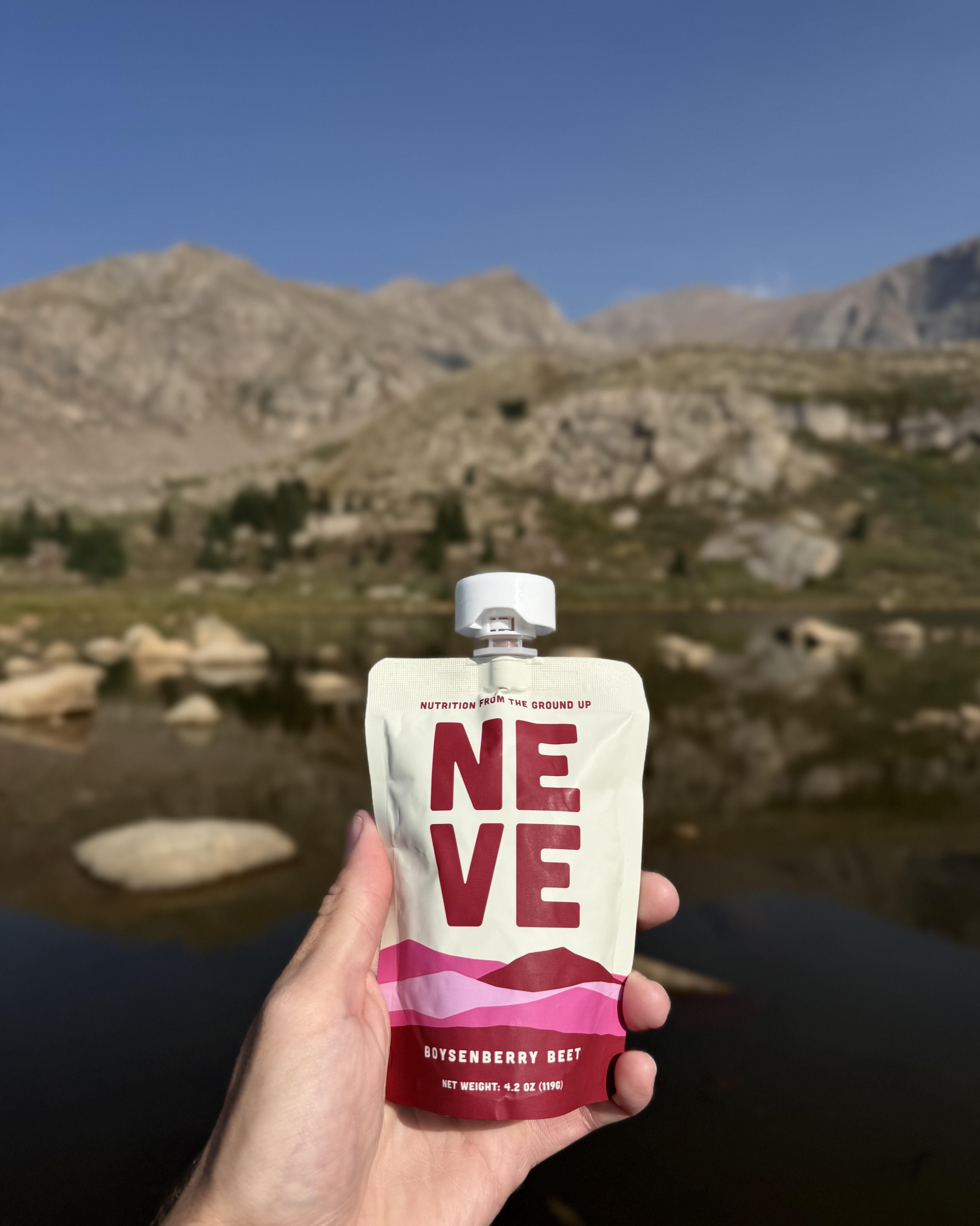 Hand holding a Neve Boysenberry Beet smoothie pouch with a mountain lake and rocky peaks in the background, duplicate file.