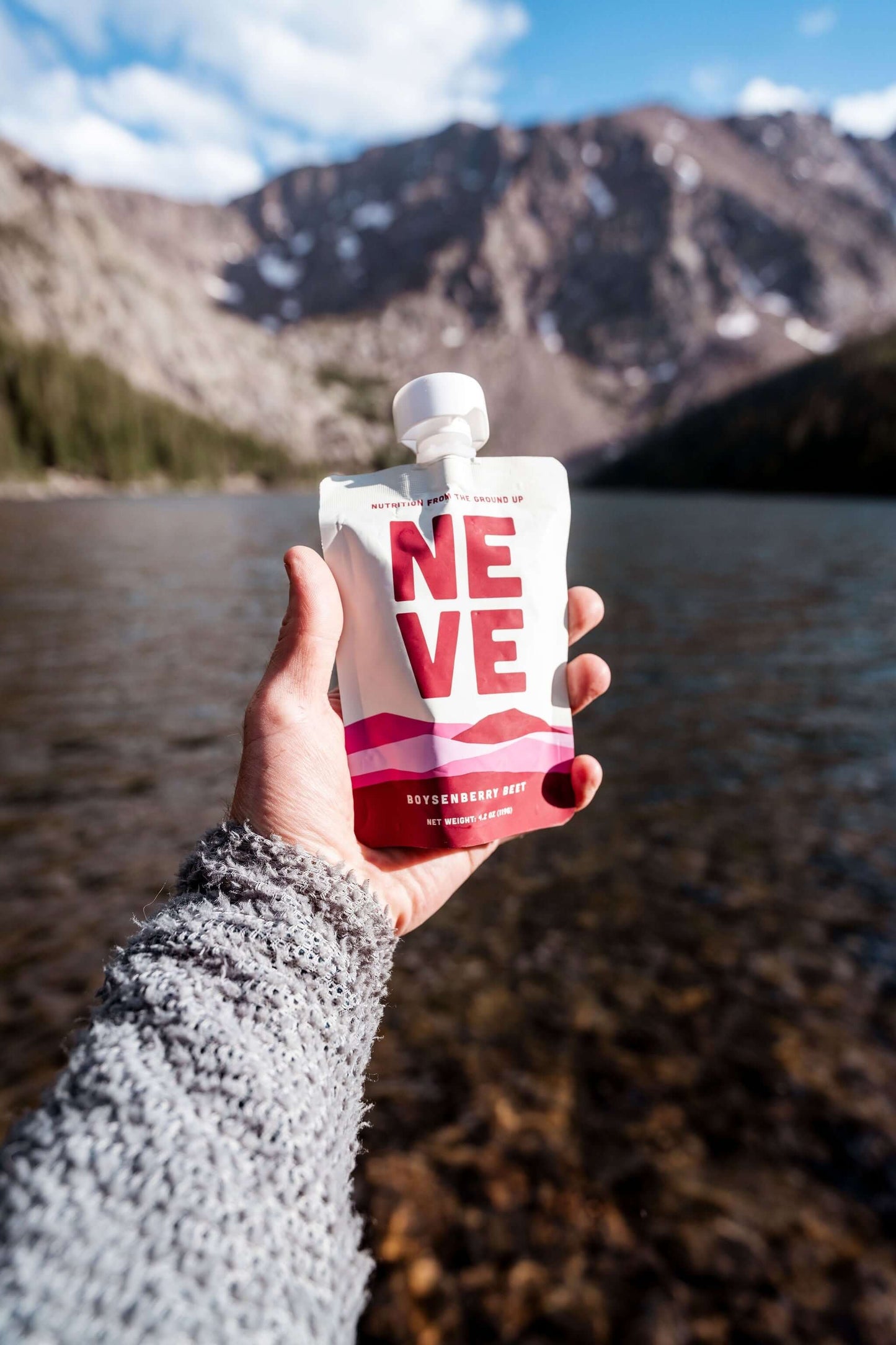 A hand in a gray knit sleeve holds a Neve Boysenberry Beet smoothie pouch in front of a scenic mountain and lake background.
