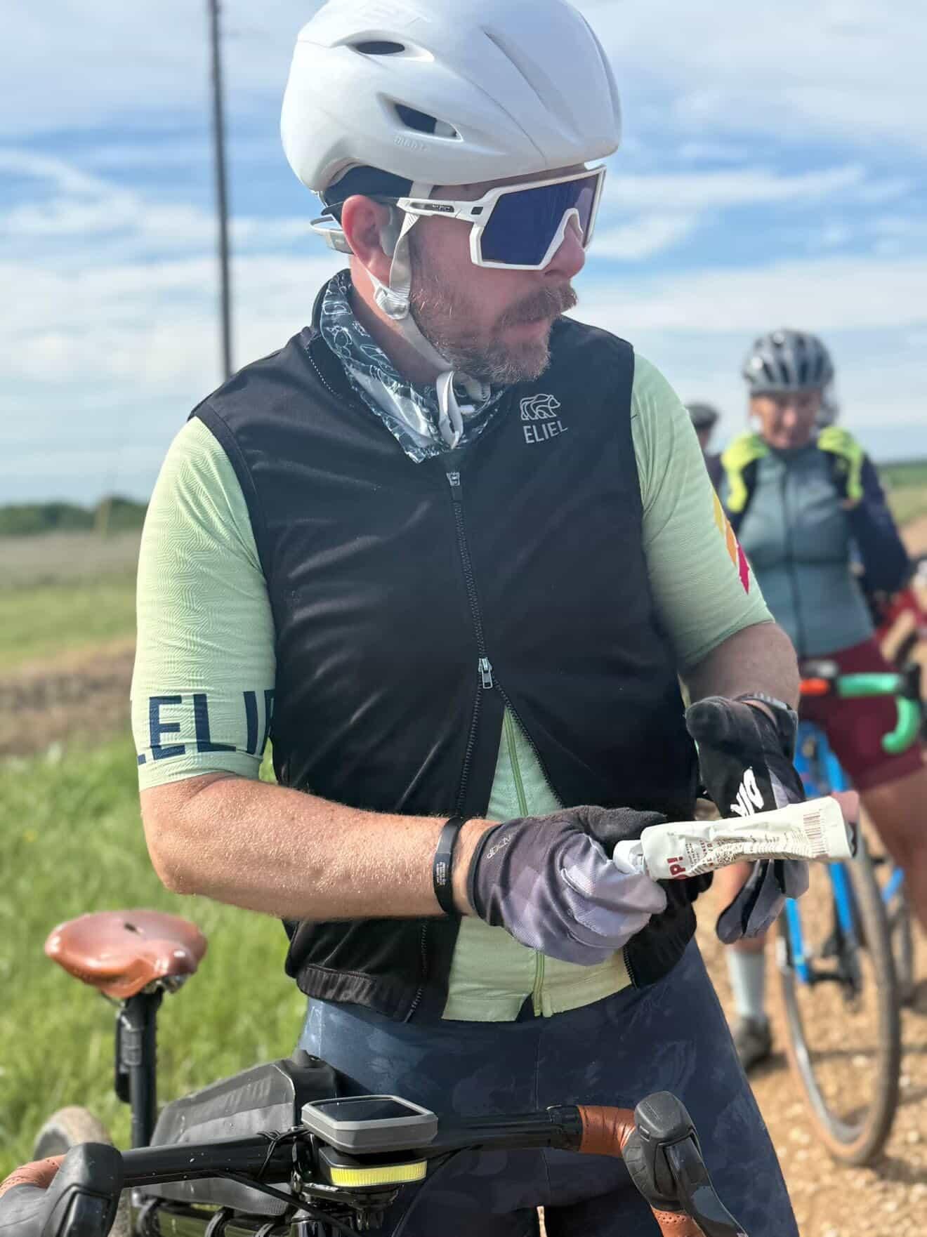 A cyclist wearing a black zippered vest over a light green jersey, a white helmet, white sunglasses, and gloves, holding an energy gel packet in front of a bicycle, with other cyclists in the background.