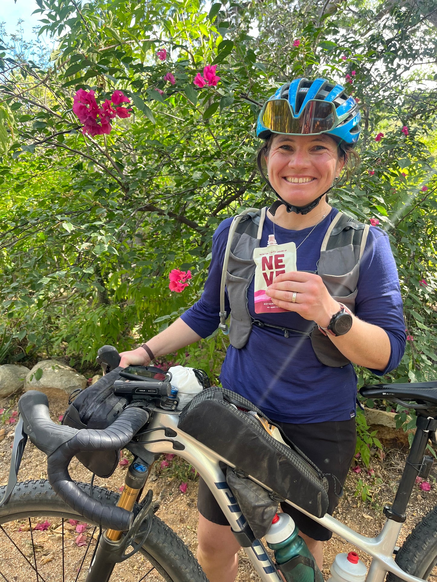 Cyclist wearing a helmet and holding a Neve smoothie pouch while standing with a bikepacking bicycle, surrounded by flowers and greenery.
