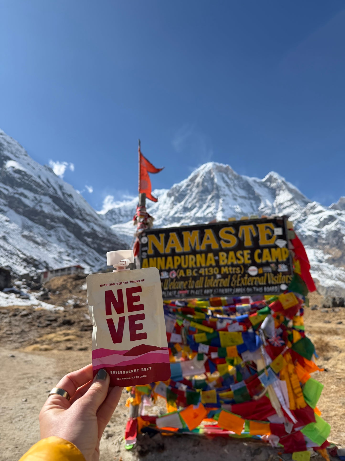 Hand holding a Neve smoothie pouch in front of the Namaste Annapurna Base Camp sign with snow-capped mountains behind.