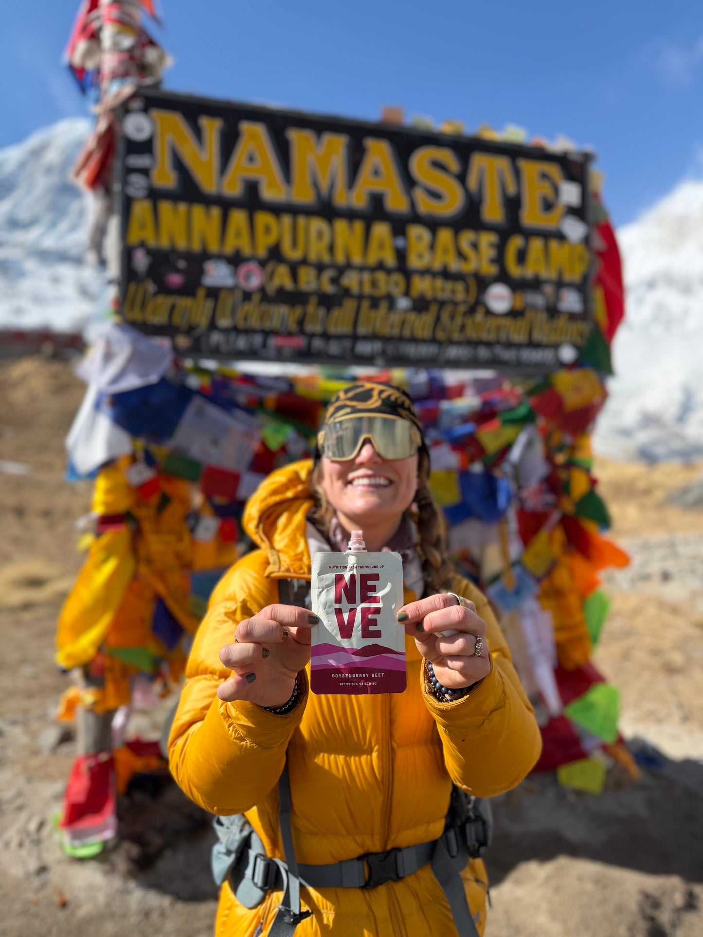 Hiker in a yellow jacket holding a Neve smoothie pouch in front of the Namaste Annapurna Base Camp sign with snowy mountains in the background.