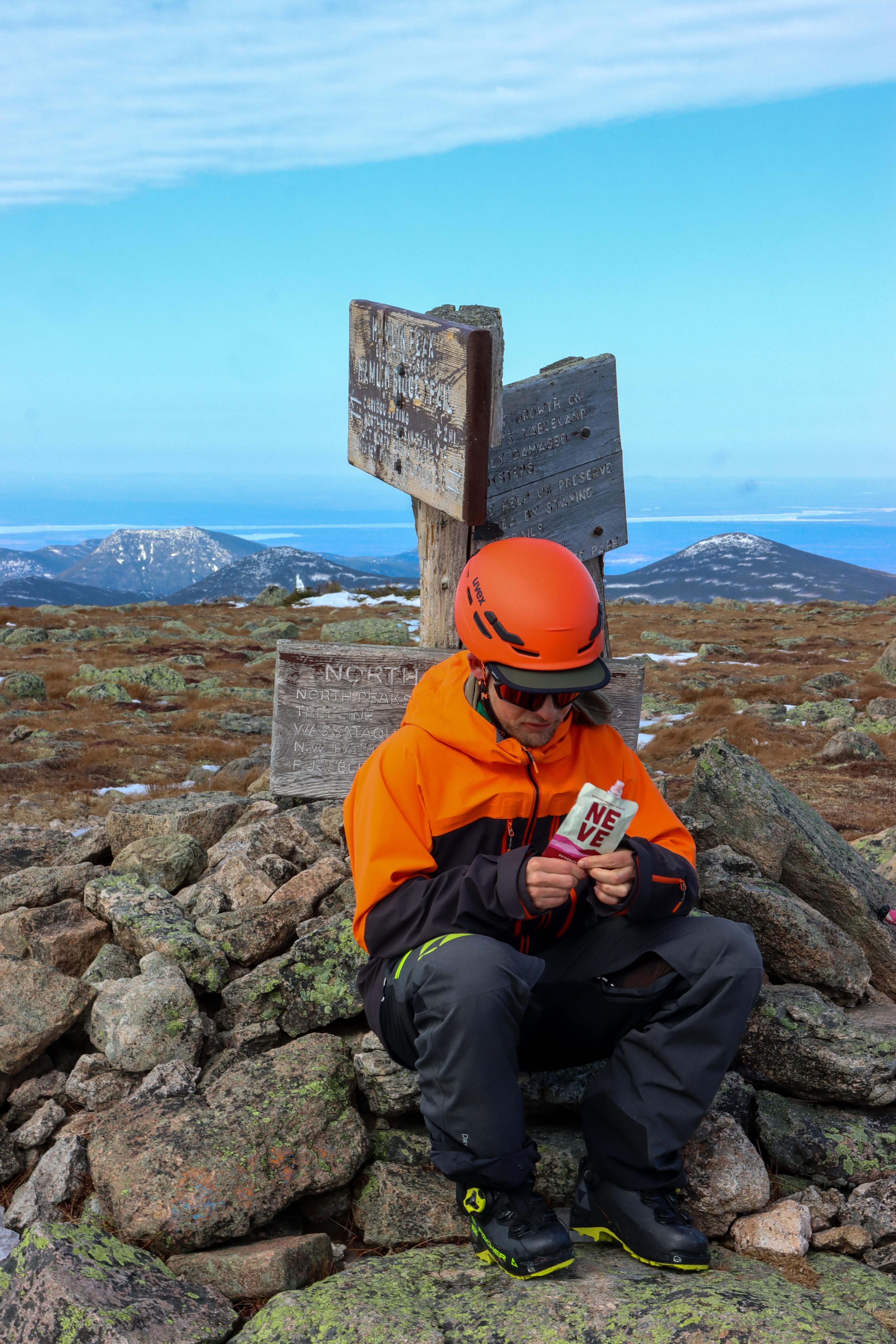 Hiker in an orange jacket and helmet sitting on rocks at a mountain summit holding a Neve smoothie pouch, with trail signs and distant peaks in the background.