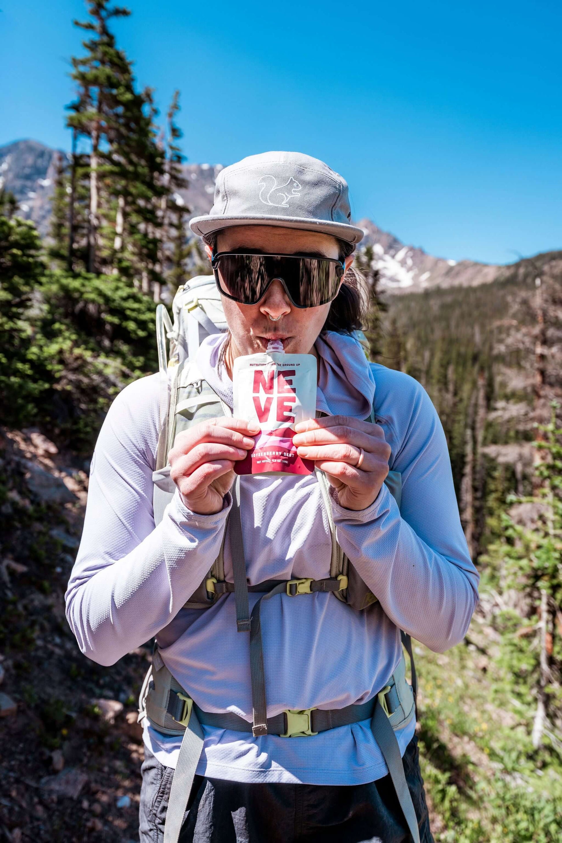 Hiker wearing a gray cap, sunglasses, and a light blue long-sleeve shirt drinking a Neve smoothie pouch, set against a mountain and pine forest background.