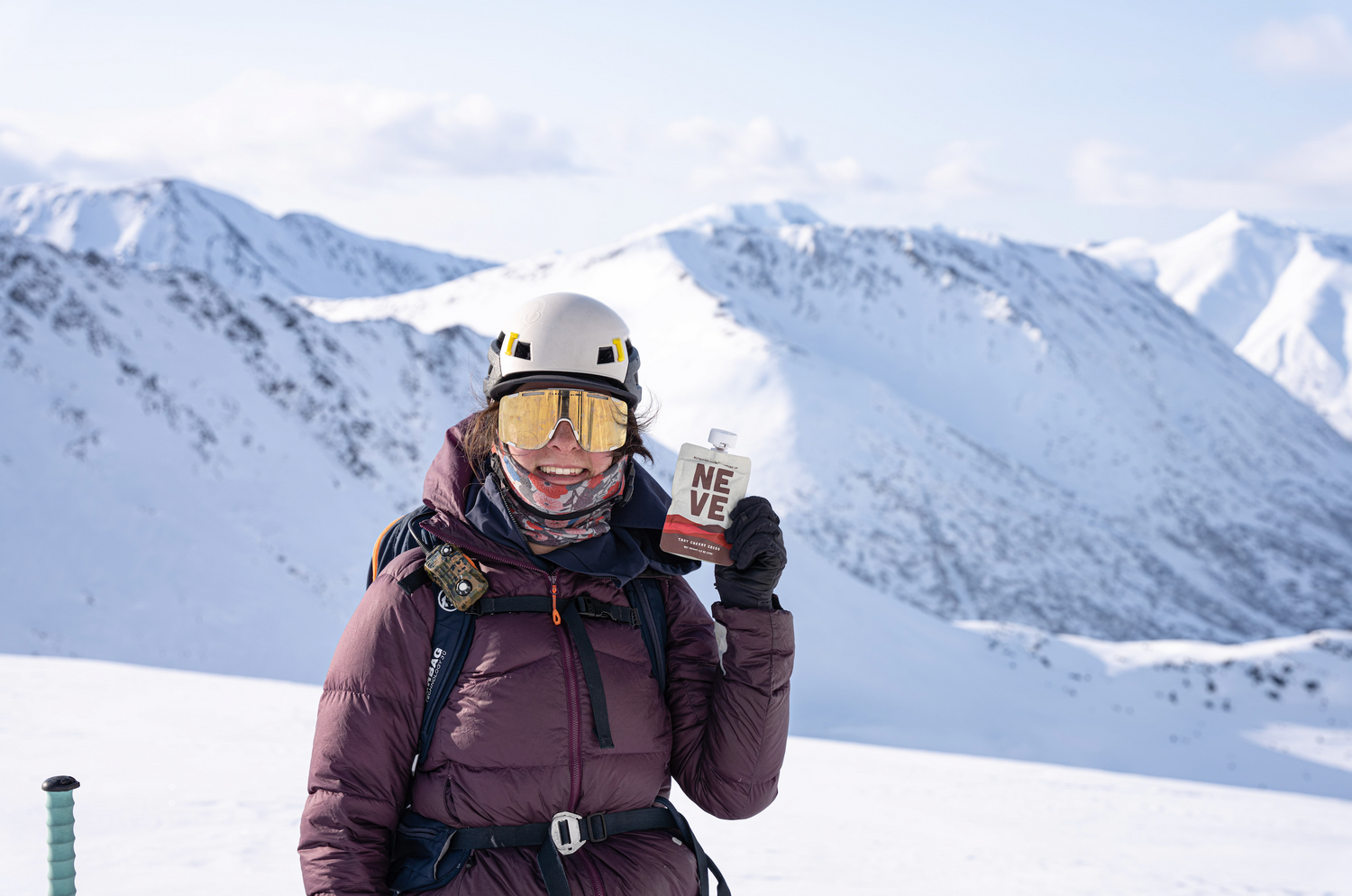 Mountaineer in winter climbing gear and helmet holding a Neve smoothie pouch, with snow-covered mountain peaks in the background.