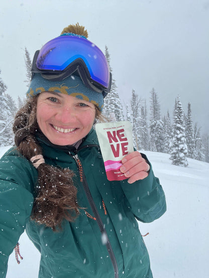 Woman in ski goggles and winter jacket taking a selfie while holding a Neve smoothie pouch, snowy forest in the background.