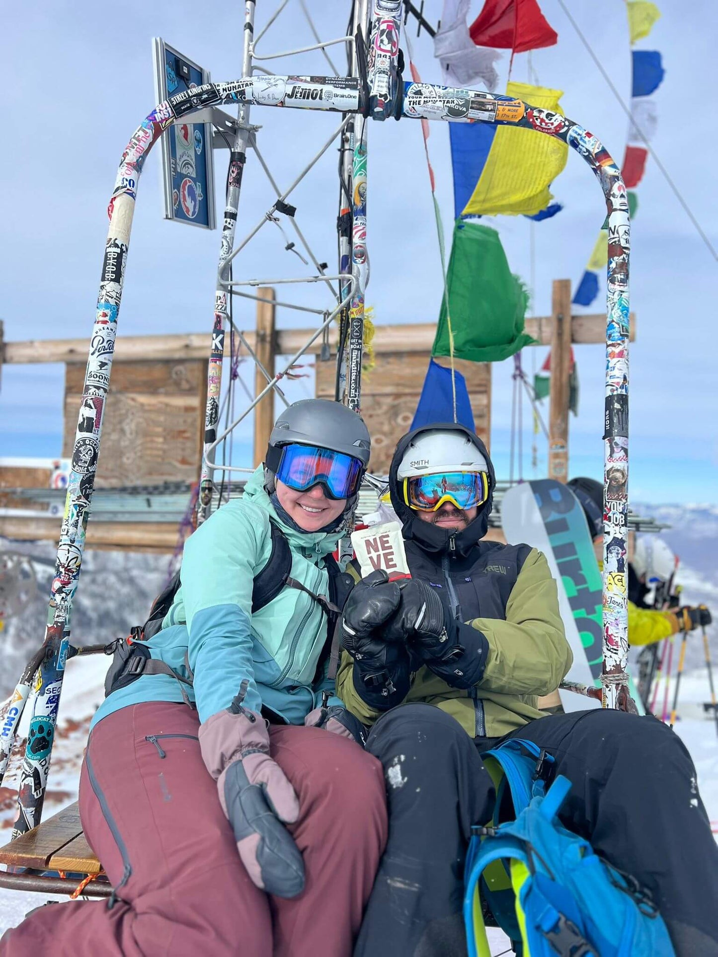 Two skiers riding a chairlift decorated with prayer flags in Aspen, one holding a Neve smoothie pouch.