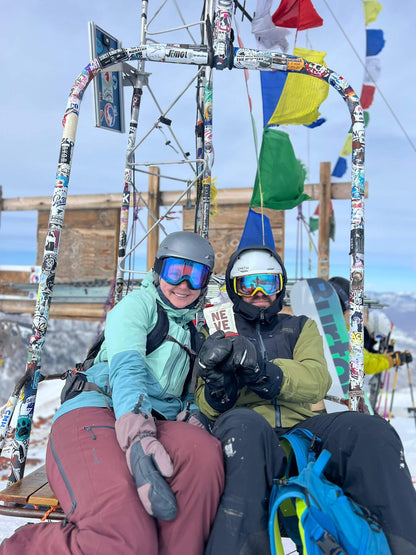 Two skiers riding a chairlift decorated with prayer flags in Aspen, one holding a Neve smoothie pouch.