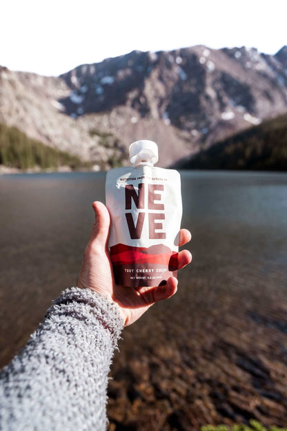 A hand in a gray knit sleeve holds a Neve Tart Cherry Cacao smoothie pouch in front of a scenic mountain and lake background.