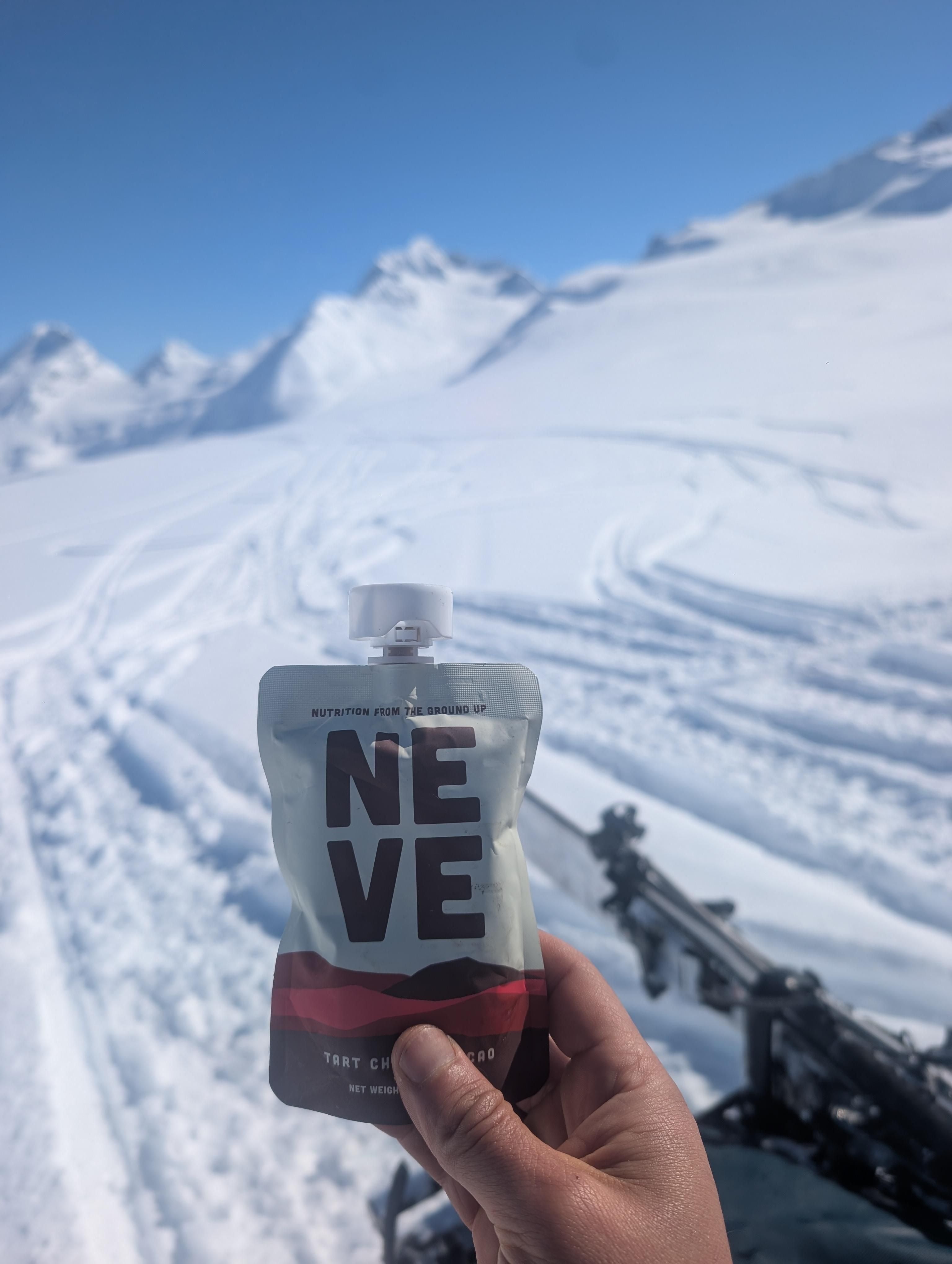 Hand holding a Neve Tart Cherry Cacao smoothie pouch with a snowy mountain slope and ski tracks in the background.